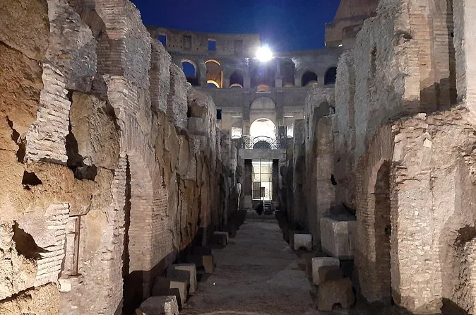 A narrow corridor in the Colosseum underground at night, lit from below, with the tiers and a bright light above